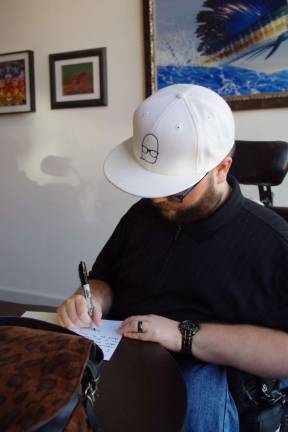 Nicholas D'Agostino is shown autographing a copy of his poetry collection book &quot;Inside Out.&quot; His distinctive logo is featured on his white ball cap.